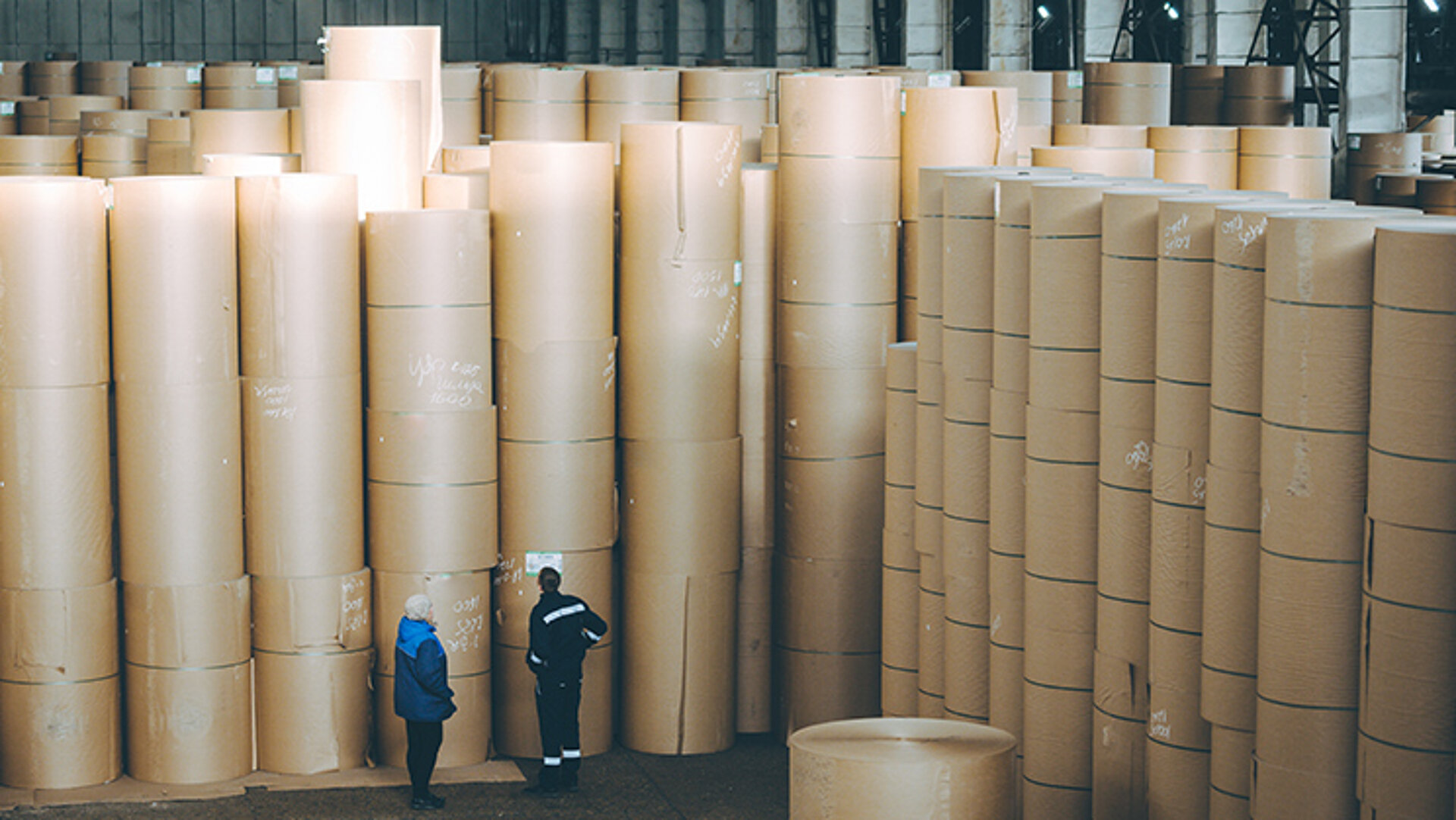 Die Lagerung von fertigen Papieren und Kartonagen erfordert speziell konstruierte Hallen. Die Produkte müssen vor Feuchtigkeit und anderen Umwelteinflüssen geschützt werden. Ist die Ware in einem Top-Zustand, kann sie versendet werden. In der Lagerhalle einer Papierfabrik befinden sich zahlreiche Stapel von bis zu sieben Rollen Wellpappenrohpapier, die von zwei Entscheidern begutachtet werden.